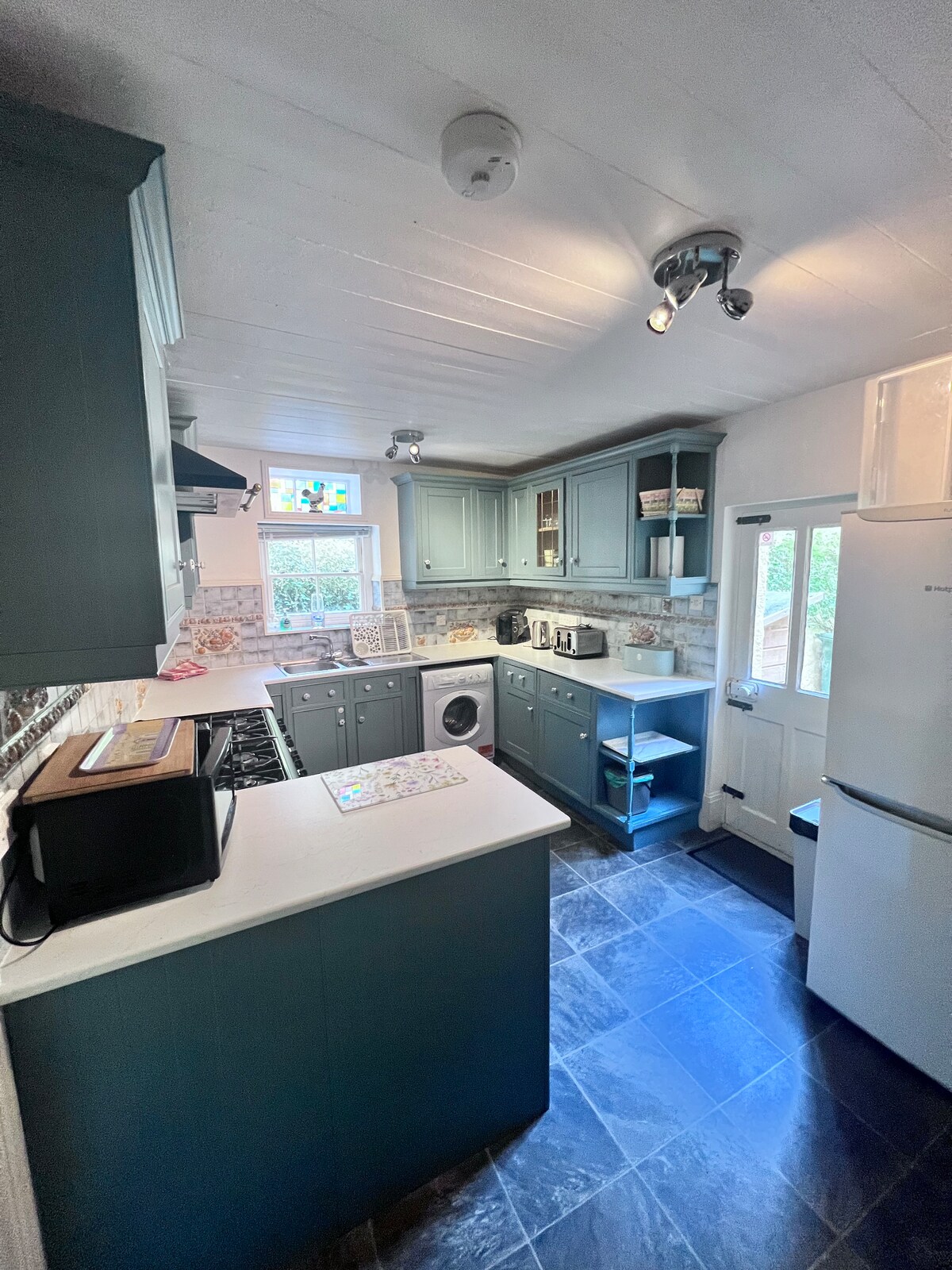The kitchen features cabinetry in a soothing blue tone, complemented by white countertops. Stainless steel appliances, including a refrigerator and oven, are present. A window allows natural light to illuminate the space, while checkerboard flooring adds a contemporary touch.