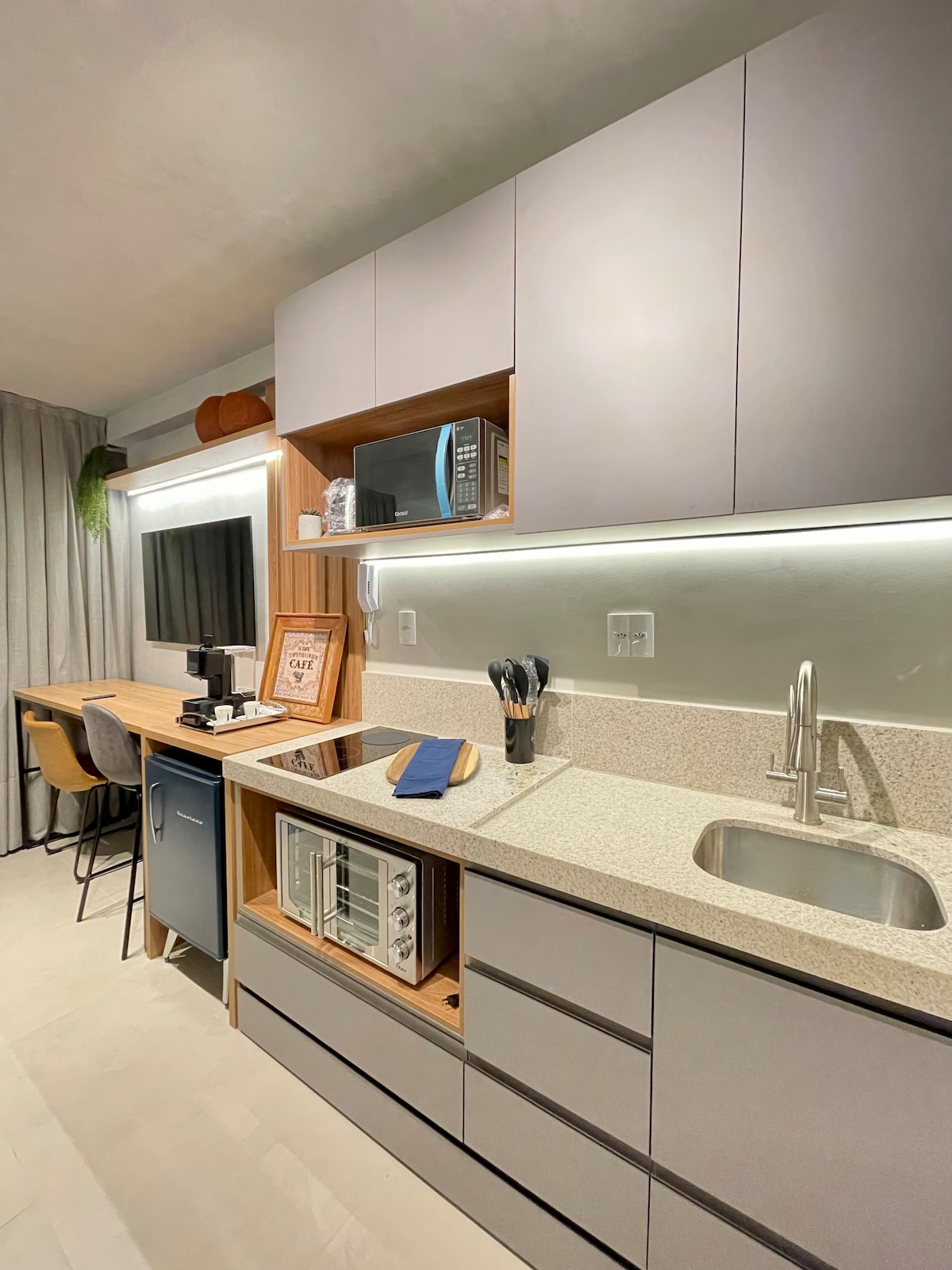 A well-equipped kitchenette features modern cabinetry and a granite countertop. Essential appliances, including a microwave and coffee maker, are arranged neatly. A workspace with seating is visible, alongside a sink and organized utensils, creating a functional area for meal preparation and dining.