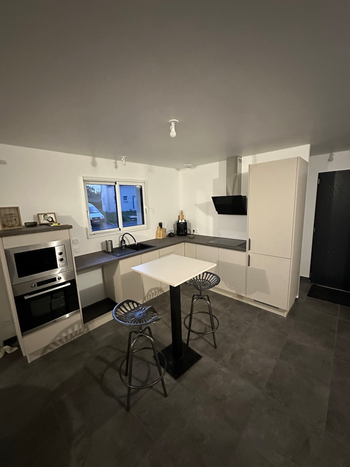 A modern kitchen is presented with sleek cabinetry and a compact dining area. A small table is paired with two bar stools, while integrated appliances, including a microwave and oven, are visible. Natural light enters through a nearby window, illuminating the tiled flooring.