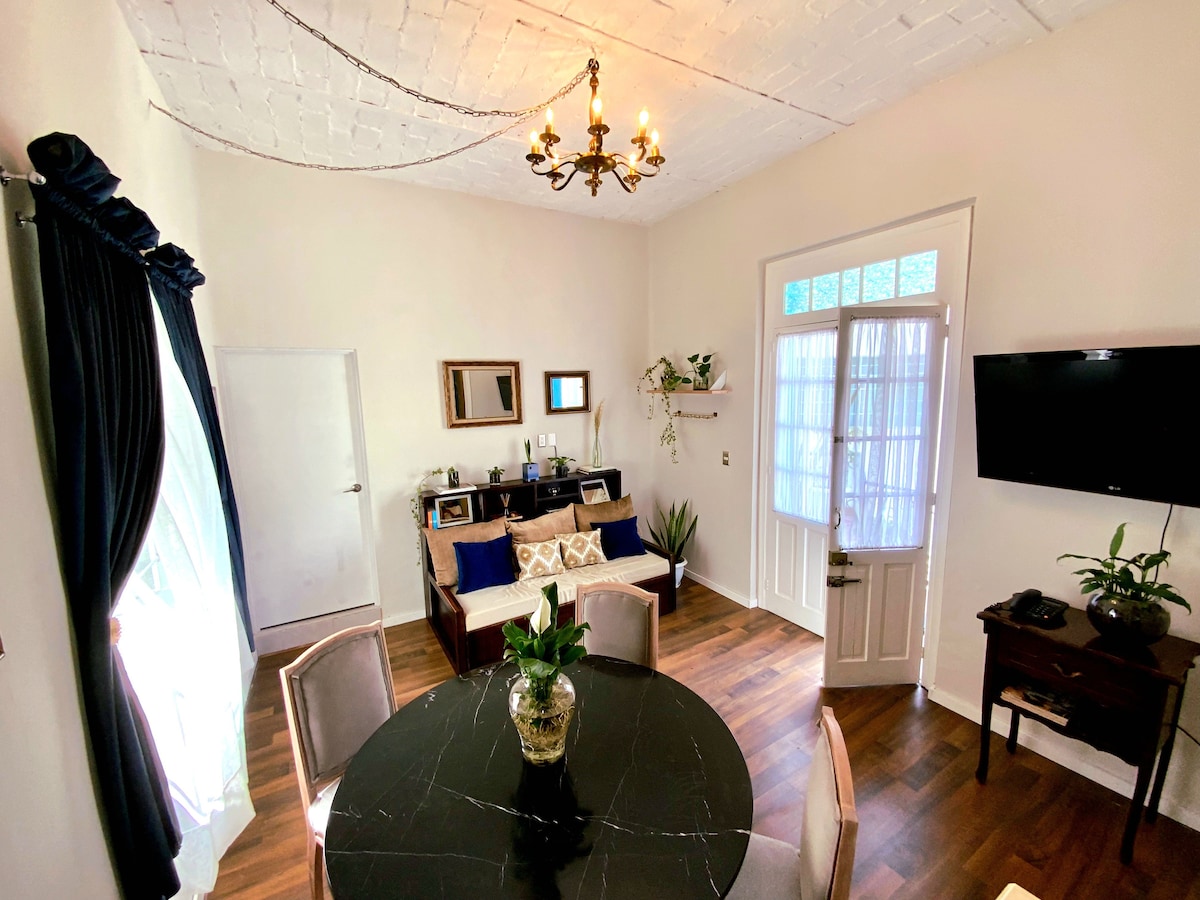 An inviting living and dining area features a round black dining table surrounded by four chairs. A comfortable sofa is positioned against the wall, and a television is mounted nearby. Natural light filters through the windows, complemented by decorative elements and a potted plant.