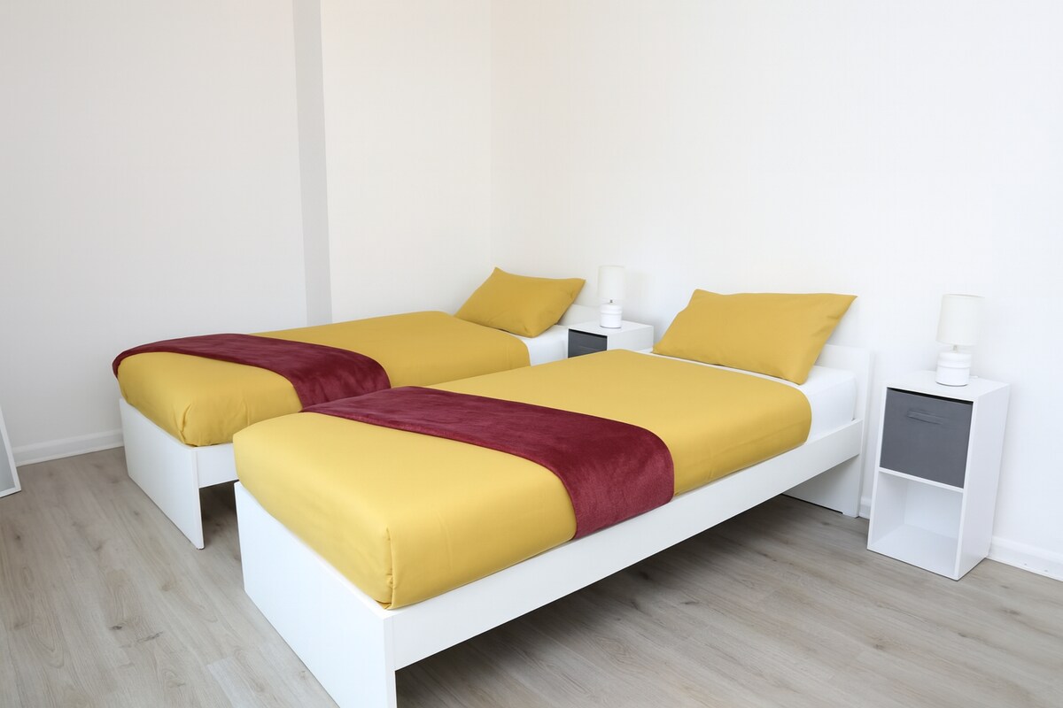 غرفة مشرقة وواسعة بيضاء بالكامل تضم سريرين مفردين مع أغطية سرير ملونة وخزانة ملابس كبيرة وخزانة ملابس وطاولات جانبية. يوفر التصميم البسيط الجديد مساحة كبيرة وراحة وتخزينًا - وهو مثالي لإقامة مريحة وعملية.