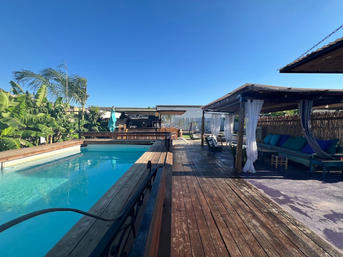 A sparkling pool is framed by a wooden deck, surrounded by tropical greenery. Comfortable seating areas with fabric drapes are positioned nearby, providing shaded relaxation spaces. A well-equipped kitchen area is visible in the background, enhancing the outdoor atmosphere.