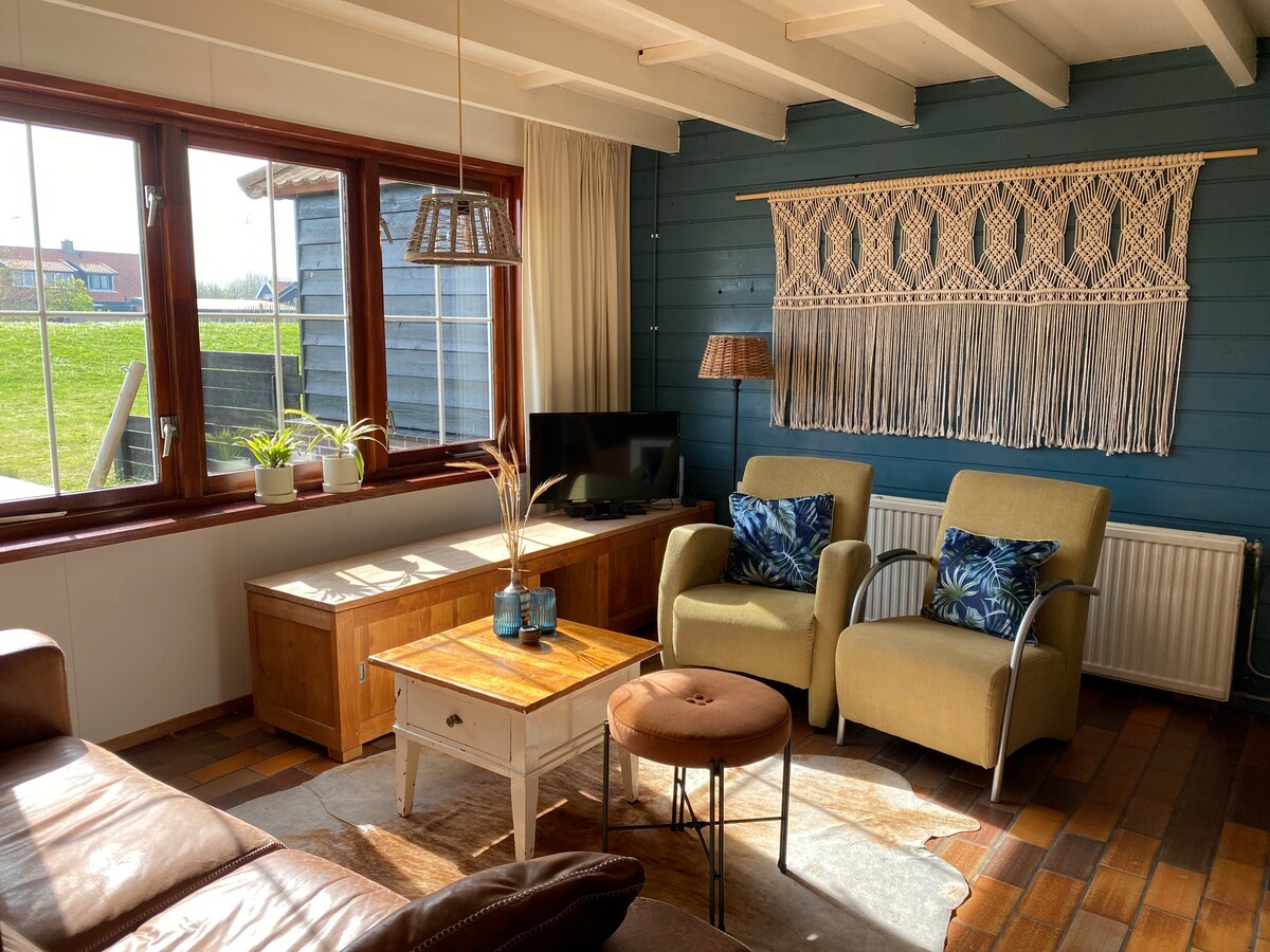 A cozy living area features large windows allowing natural light to fill the space. Two green armchairs are positioned near a wooden coffee table, with a woven wall hanging above. A television is placed on a stand, complemented by potted plants on the window sill.