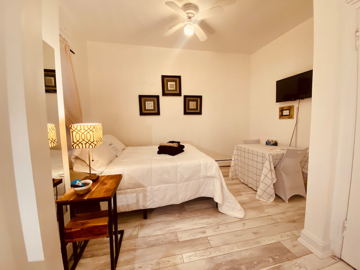 A bright room features a spacious bed dressed in white linens, complemented by decorative wall frames. A small table with a lamp stands next to the bed, and a television is mounted on the wall. Light-colored flooring enhances the airy feel of the space.