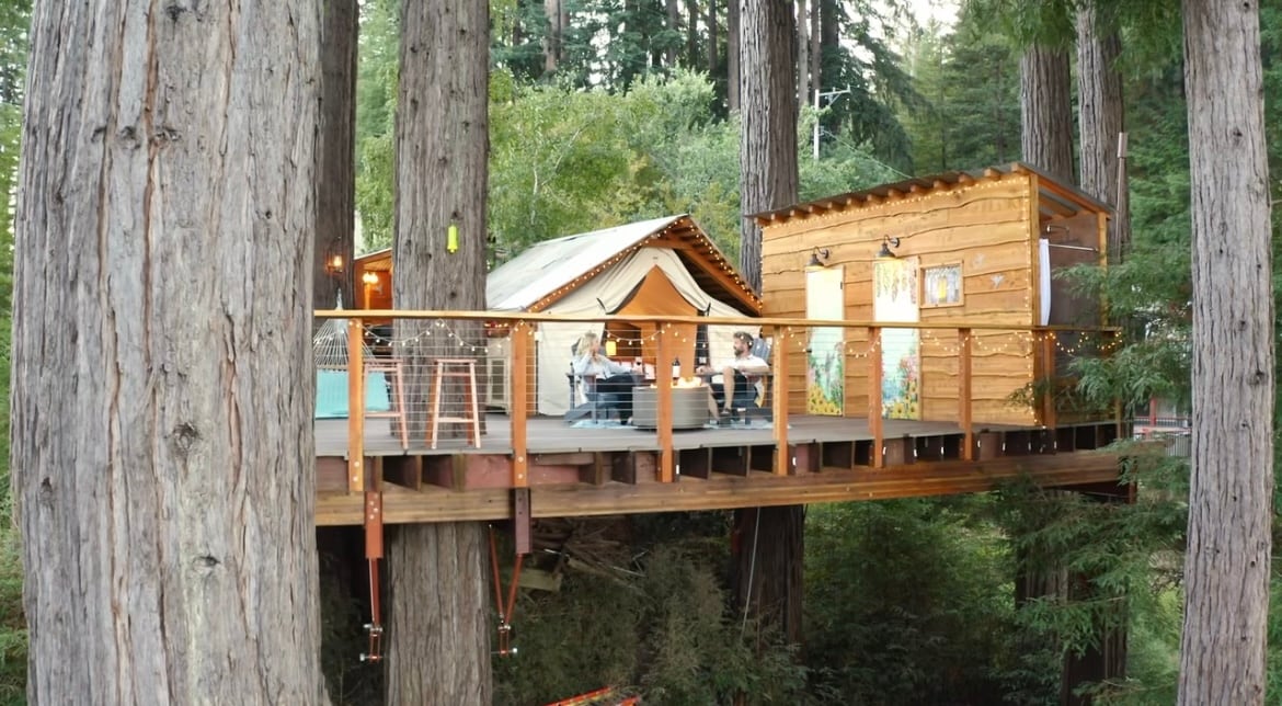 A tranquil glamping retreat is perched among towering redwoods, featuring a canvas tent and a cozy wooden structure. A spacious deck is adorned with soft lighting, offering a serene space for relaxation and gatherings in nature.