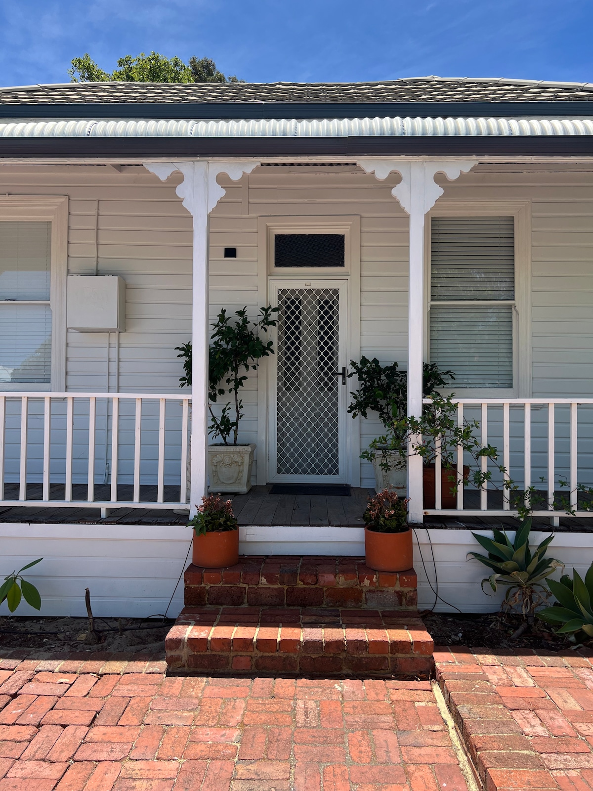 The front entrance of the heritage cottage is framed by a white porch with decorative railings. Potted plants are arranged along the steps leading up to a screen door, complemented by a brick pathway and vibrant greenery.