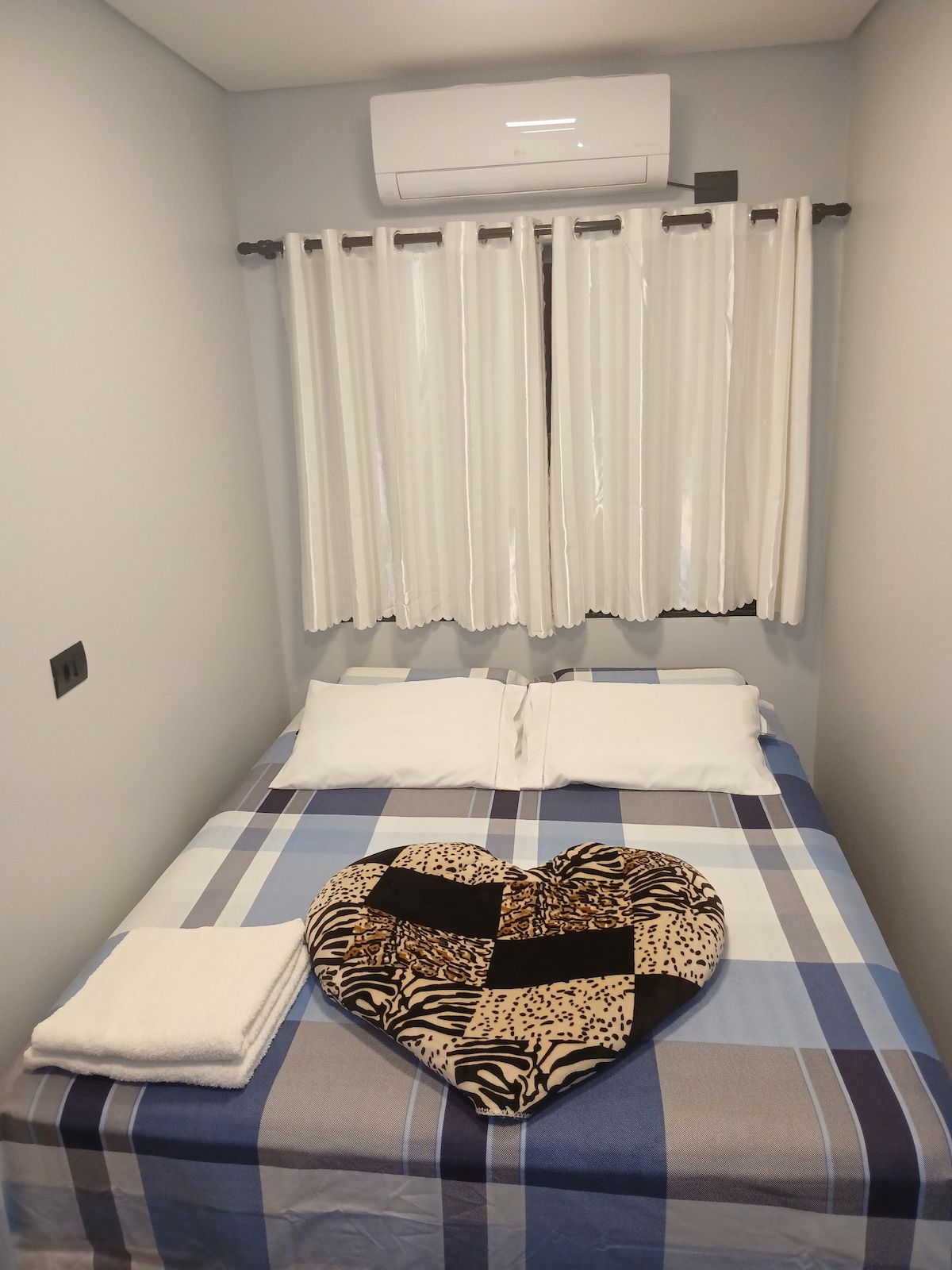 A cozy sleeping area features a spacious bed adorned with a heart-shaped decorative pillow against a plaid blanket. Two pillows rest at the head of the bed, and light filters softly through sheer curtains covering the window.