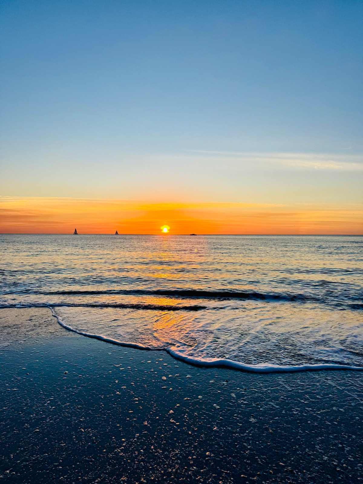 A serene beach scene captures the sunset on the horizon, where the sun's warm glow reflects on gentle ocean waves. Silhouettes of distant sailboats can be seen against the colorful sky transitioning from orange to blue.