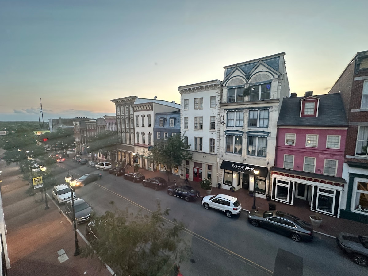 A view of a vibrant street in Wilmington showcases a mix of architectural styles from historic to contemporary. Various vehicles are parked along the curb, and trees add greenery to the scene. The setting sun casts a warm glow over the buildings.