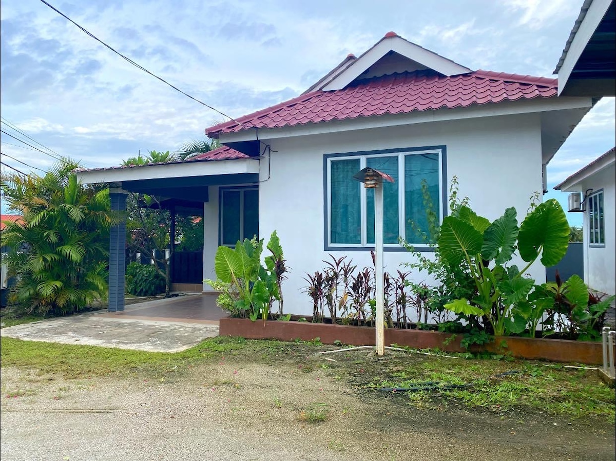 A single-story home is showcased with a spacious porch and a red-tiled roof. Surrounding the entrance are neatly arranged greenery and small plants, adding a touch of nature. A paved walkway leads to the front door, enhancing accessibility.