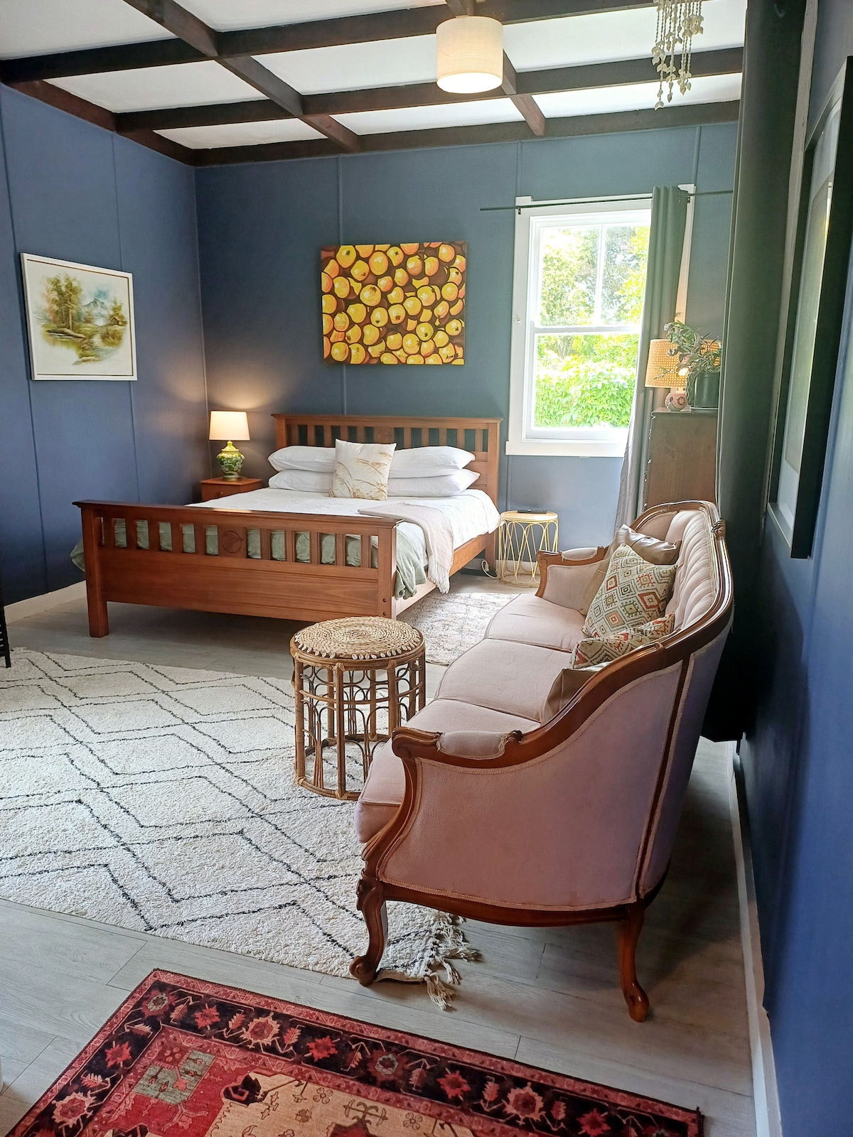 A cozy bedroom features a wooden bed with white linens and decorative pillows. A pink settee with patterned cushions complements the space. Natural light is allowed in through a window surrounded by dark curtains, while a textured rug adds warmth to the light-colored flooring.