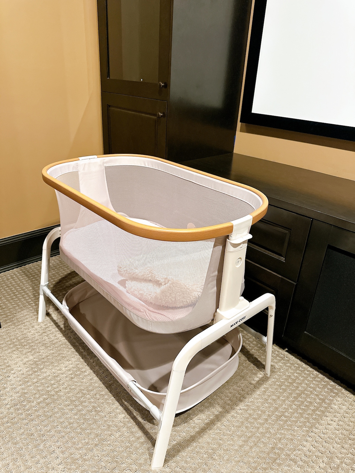 A modern bassinet is positioned beside a dark wooden cabinet, featuring a soft mesh side that provides visibility. The bassinet has a wooden rail around the top and includes a storage area beneath. The room is softly illuminated, contributing to a calm environment.