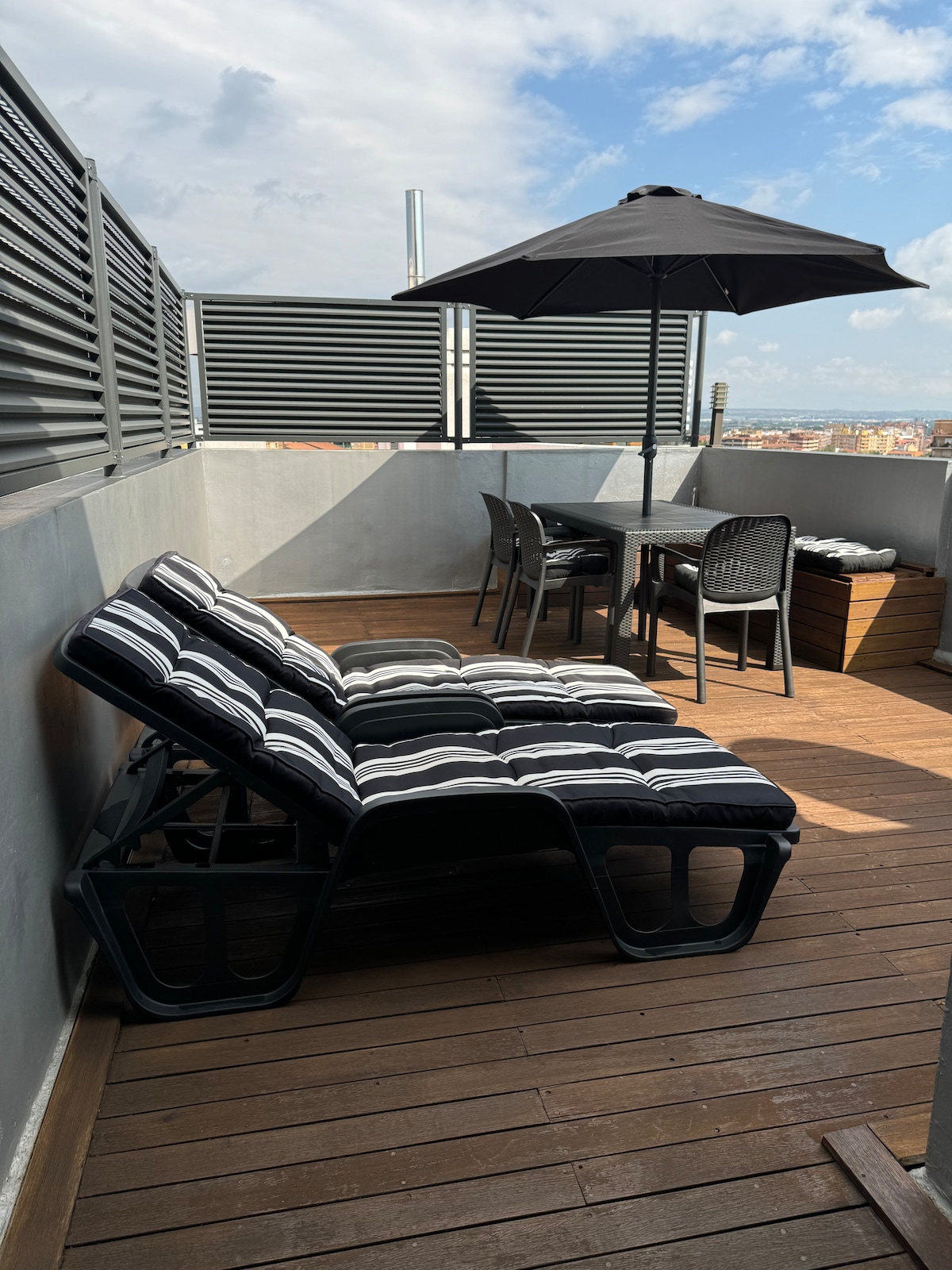A spacious terrace is furnished with a striped sun lounger, a dining table for four, and a black umbrella for shade. Wooden decking surrounds the area, complemented by modern privacy screens that enhance the design. Views of the skyline are visible in the background.