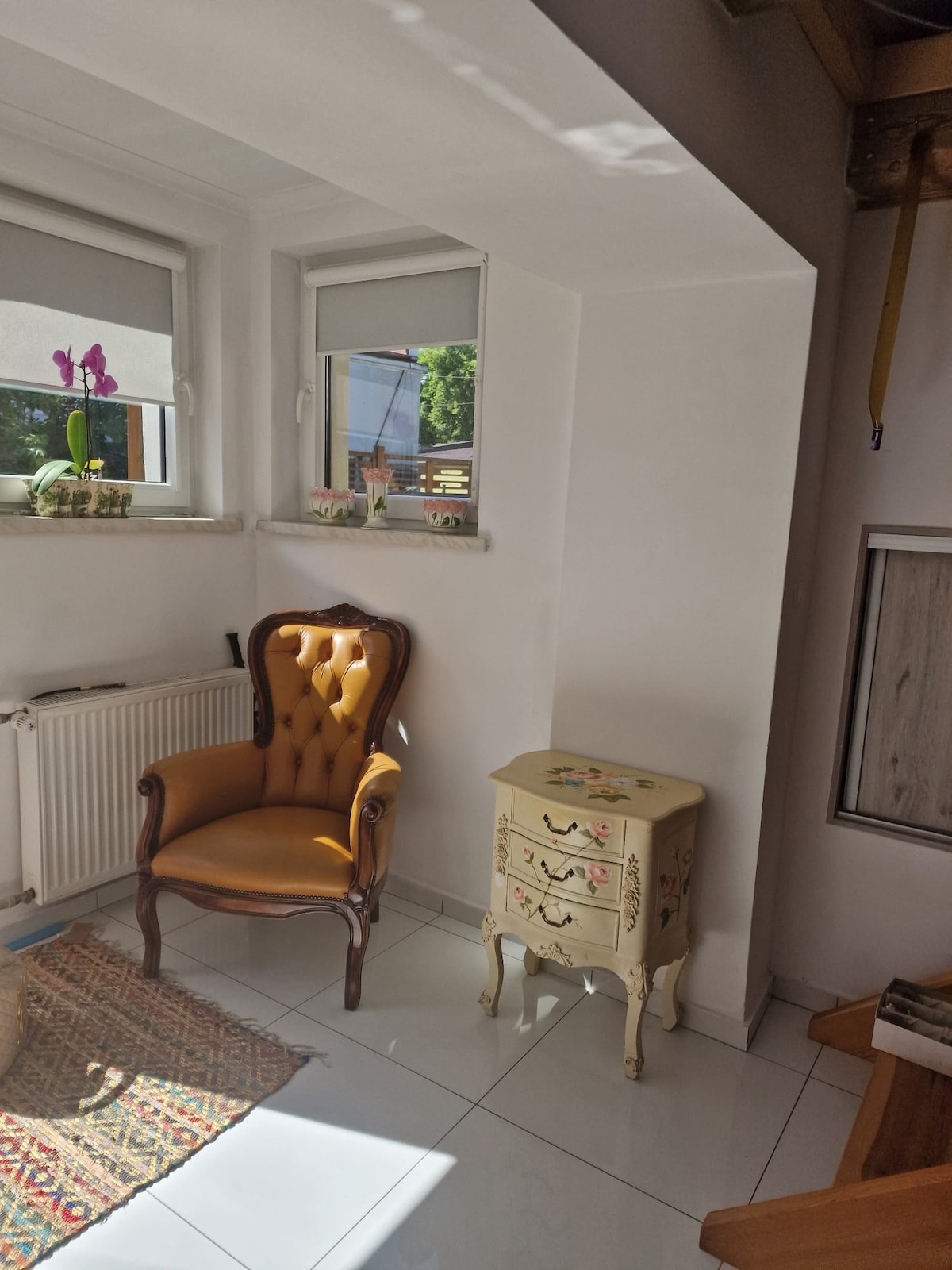 A bright corner features a vintage armchair with button tufting alongside a small, intricately painted dresser. Natural light floods the space through two windows, illuminating decorative plants on the windowsill and highlighting the glossy white tiles and minimalist decor.