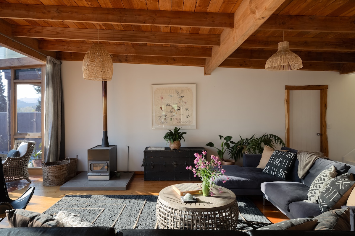 A spacious living area features a comfortable sectional sofa and a wooden coffee table adorned with fresh flowers. A modern log burner is centered on a wood-paneled wall, complemented by decorative plants. Natural light brightens the room, showcasing its welcoming atmosphere.