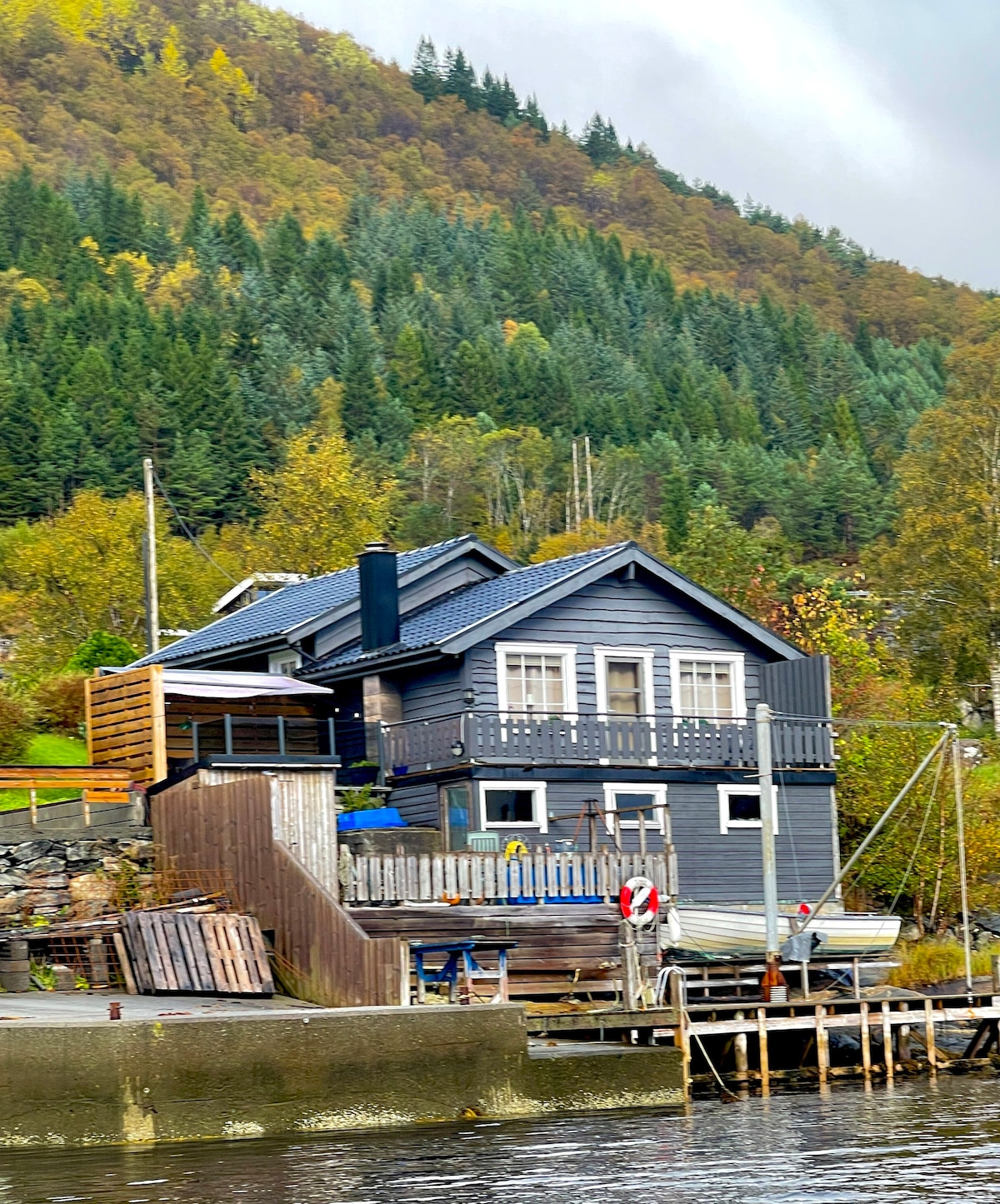 A two-story wooden house is positioned near the water, featuring a dark exterior with light-colored windows. A balcony extends from the upper level, providing views of the surrounding greenery. A wooden walkway leads to a dock with a small boat at the water's edge.