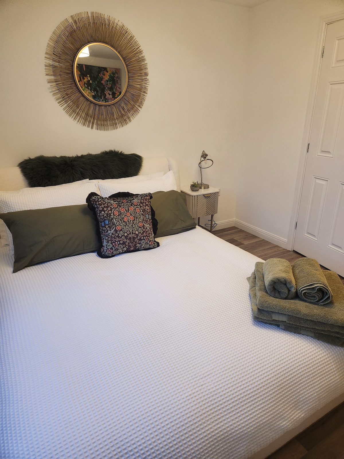 A clean and recently decorated bedroom features a spacious bed topped with decorative pillows. Soft towels are neatly arranged on the edge, and a small bedside table holds a lamp. A round mirror with a natural design is mounted above the headboard.