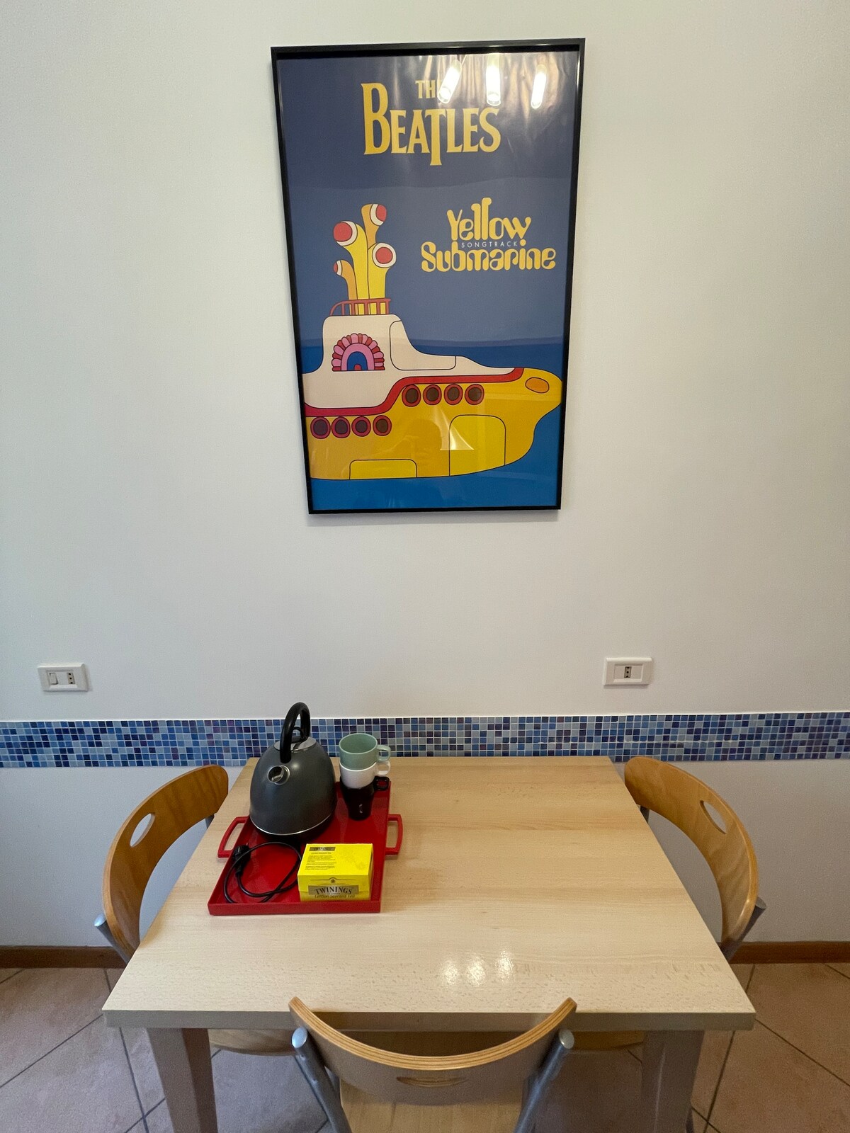 A dining corner is presented with a light wooden table and two chairs. A vibrant poster featuring a colorful design hangs above, while a kettle, cups, and a tray with items are neatly arranged on the table. The walls are accented with a mosaic border.