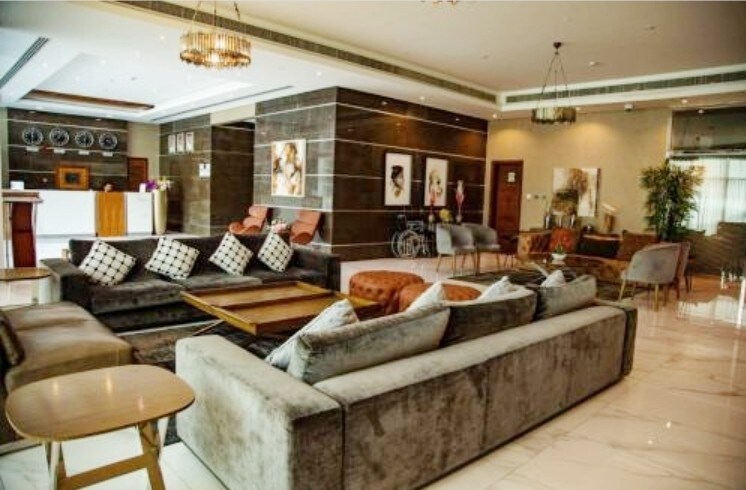 The lobby area features a modern design with a mix of comfortable seating options, including plush sofas and chairs arranged around a central coffee table. Artwork adorns the walls, and a reception desk is visible in the background, enhancing the welcoming atmosphere.