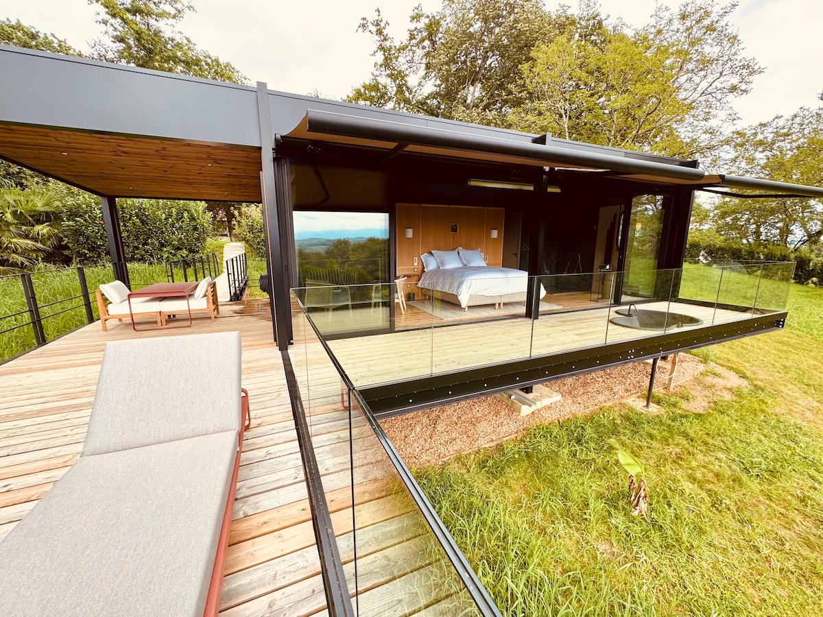 A contemporary suite is showcased with large glass walls, allowing for natural light and stunning views. An outdoor terrace features lounge seating and a pathway leading to lush greenery. The interior includes a spacious bed and modern décor, highlighting the seamless connection between inside and outside.