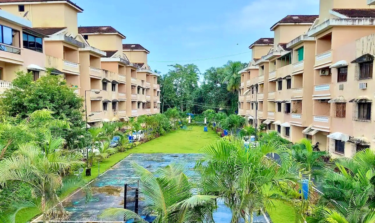 A landscaped courtyard is framed by two apartment buildings, surrounded by lush greenery and palm trees. A clear blue sky enhances the serene atmosphere, while a peaceful garden pathway leads through the area. A glimpse of a recreational space is visible on the left.