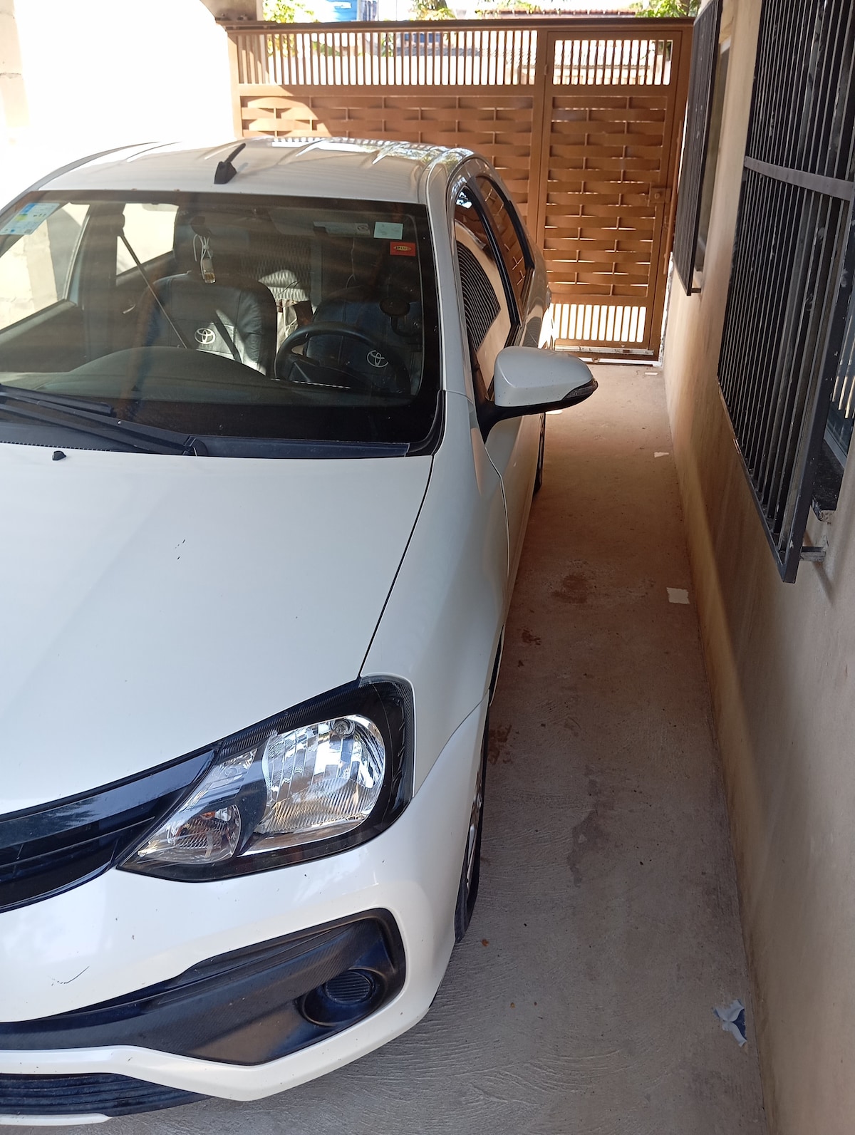 A white car is parked in a narrow driveway, flanked by walls on either side. The vehicle is positioned close to a wooden gate, partially open, revealing a light-colored exterior wall. Illuminated by natural light, the surface shows a smooth finish.
