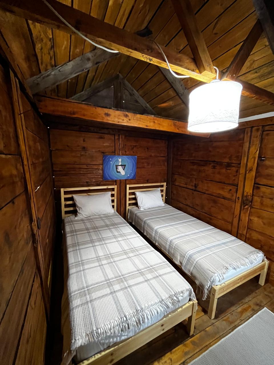 A cozy sleeping area features two wooden single beds, each dressed in light grey blankets. The room showcases wooden walls and beams, creating a rustic ambiance. A shaded light fixture hangs from the ceiling, providing soft illumination, while a piece of blue wall art adds a pop of color.