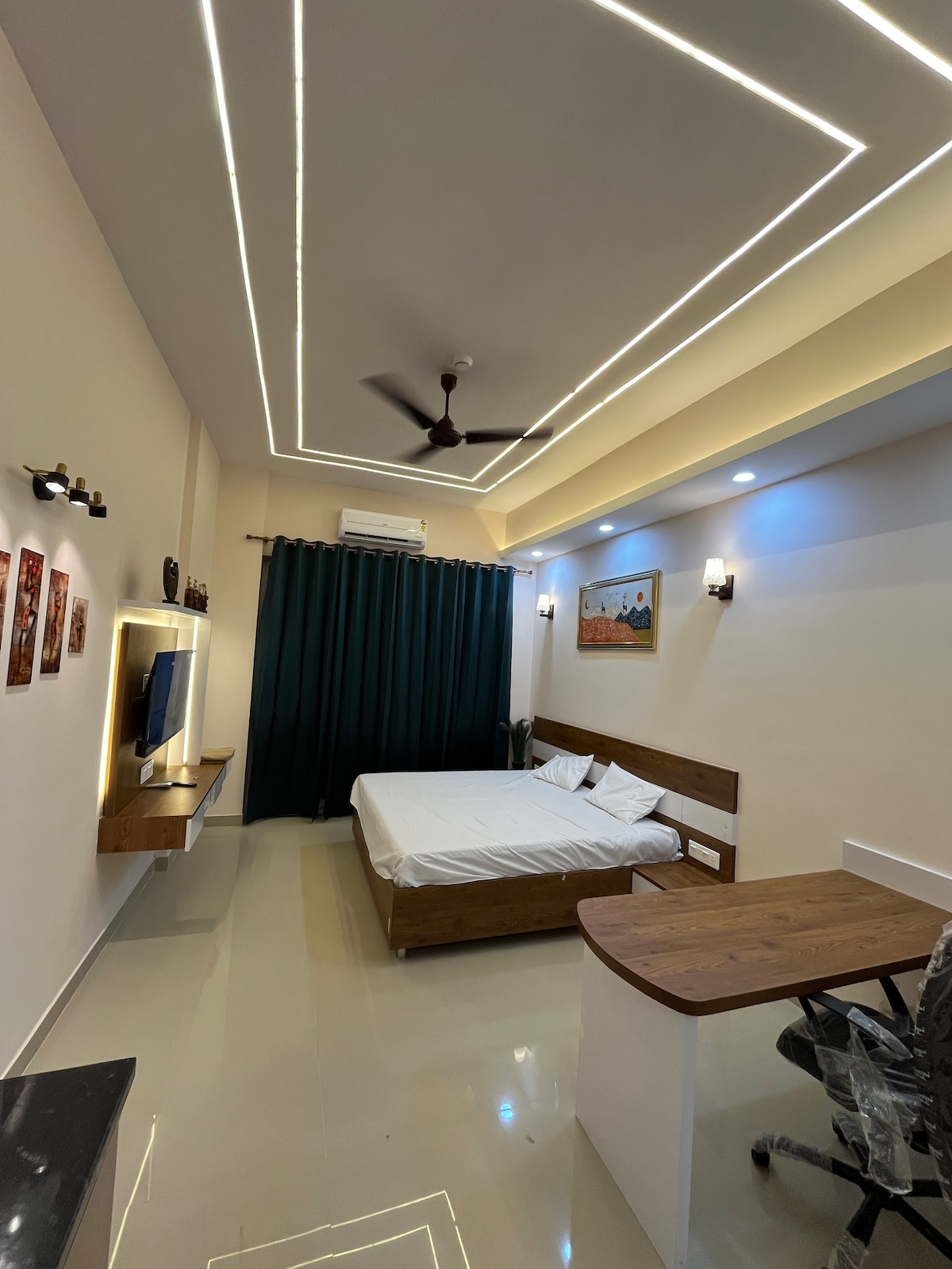 A compact studio apartment features a spacious bed with white linens against a neutral backdrop. A work desk and chair are positioned nearby, while a TV is mounted on the wall. Large curtains frame a window, and modern lighting highlights the ceiling design.