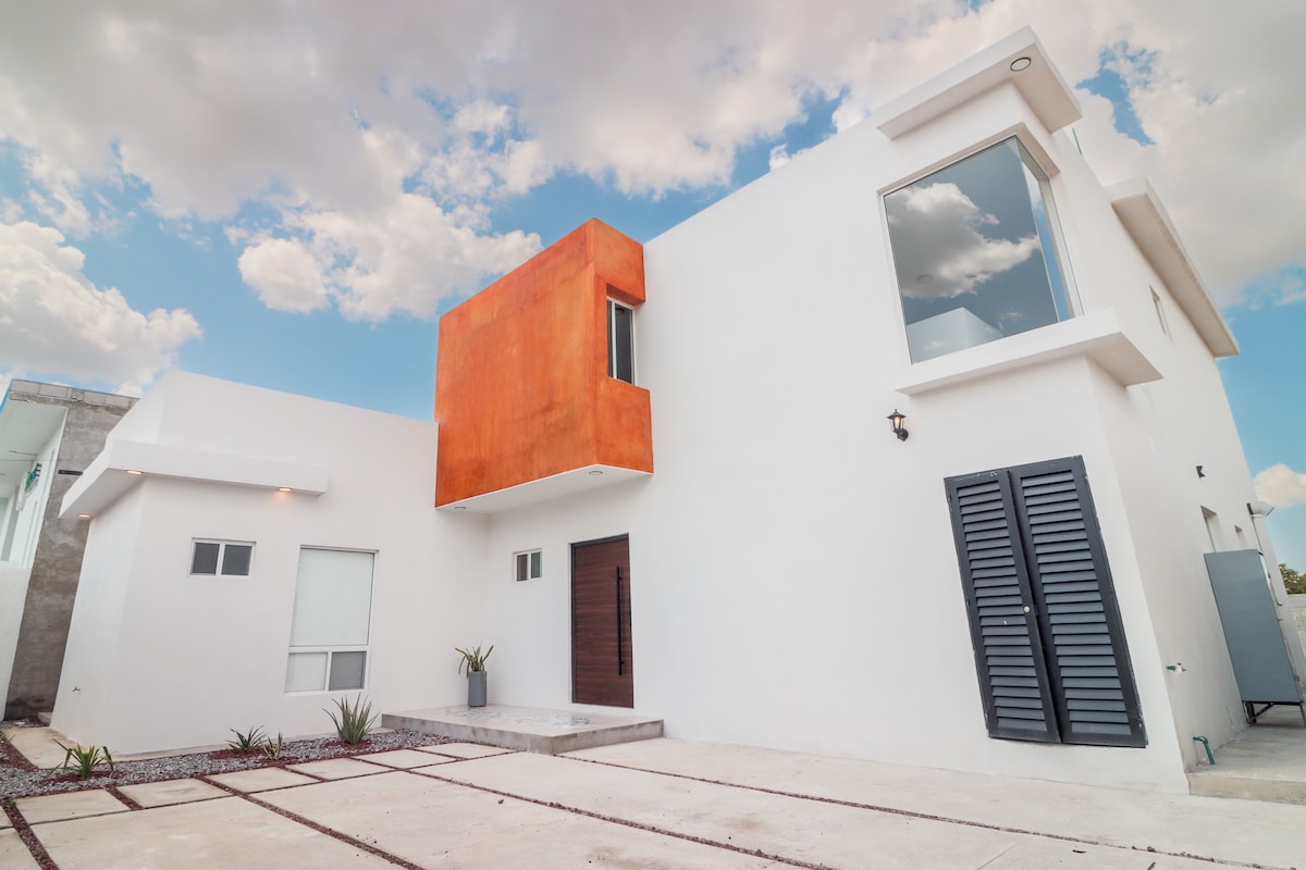 A modern two-story house is presented with a white exterior and an accent orange wall. Large windows allow natural light to illuminate the interior. A simple pathway leads to the entrance, which features a wooden door flanked by a small decorative plant.