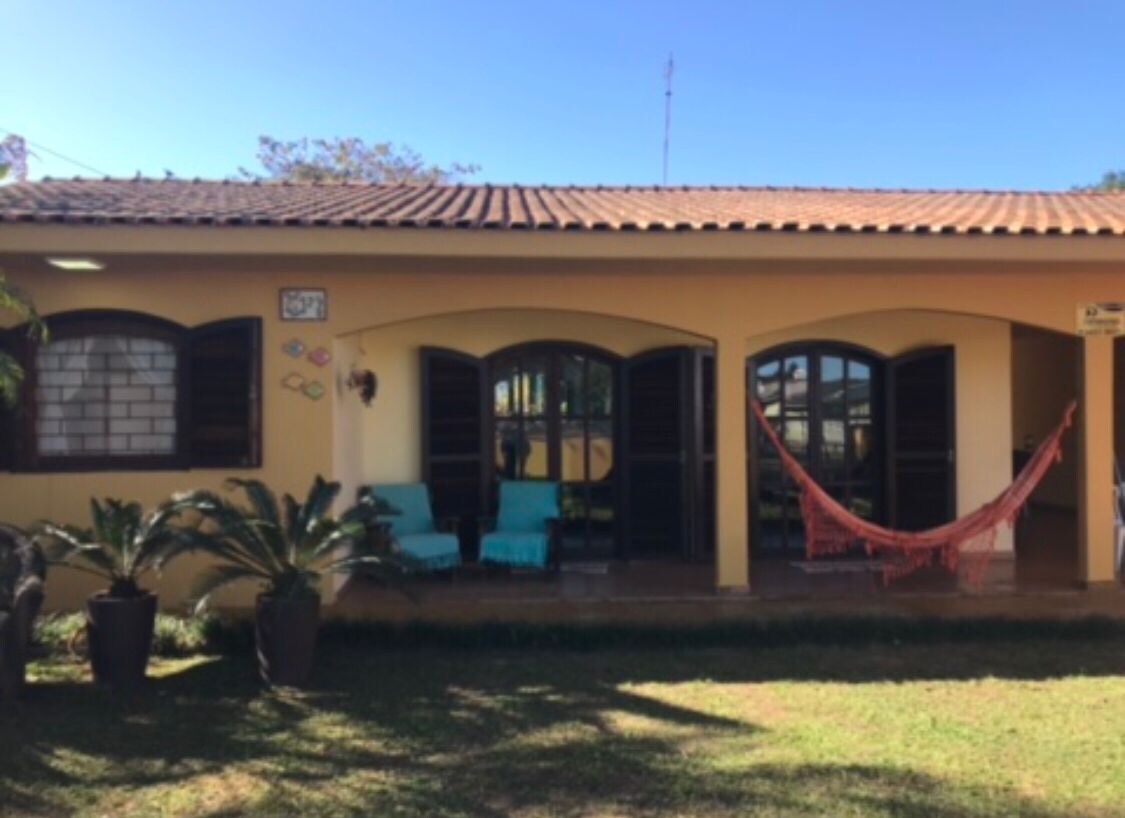 A single-story house is featured with a warm-toned exterior and a tiled roof. Two bright blue chairs are positioned on the porch, complemented by a colorful hammock hanging between two columns. Lush greenery surrounds the entrance, enhancing the inviting outdoor space.