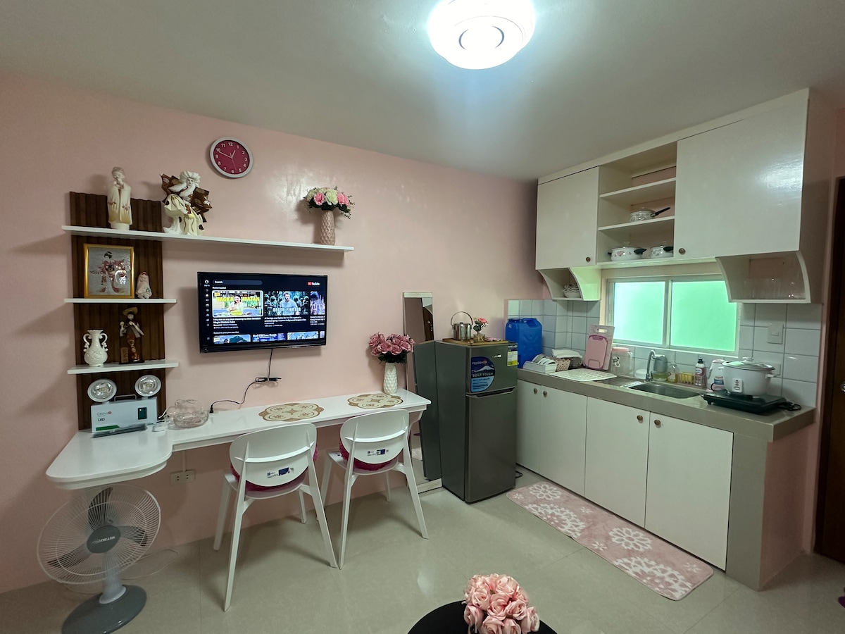The kitchen area features pink walls and a combination of white cabinetry and appliances. A dining table with three chairs is placed near a wall-mounted TV. A refrigerator and cooking appliances are visible, with a window allowing natural light to enter the space.