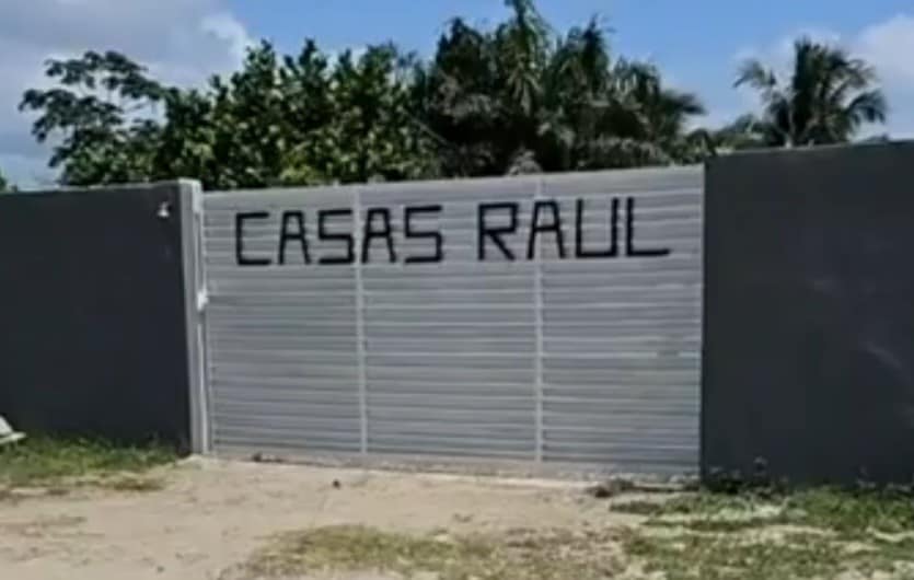 The entrance to Casas Raul is marked by a large gray gate with the name 'CASAS RAUL' prominently displayed in bold letters. Lush green trees are visible in the background under a clear blue sky.