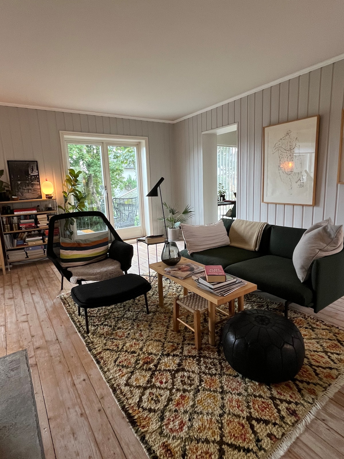 A comfortable living area features a mix of seating options, including a green sofa and a stylish black chair. A wooden coffee table holds various books and a decorative vase. Natural light filters through large windows, illuminating the wooden floor and patterned area rug.