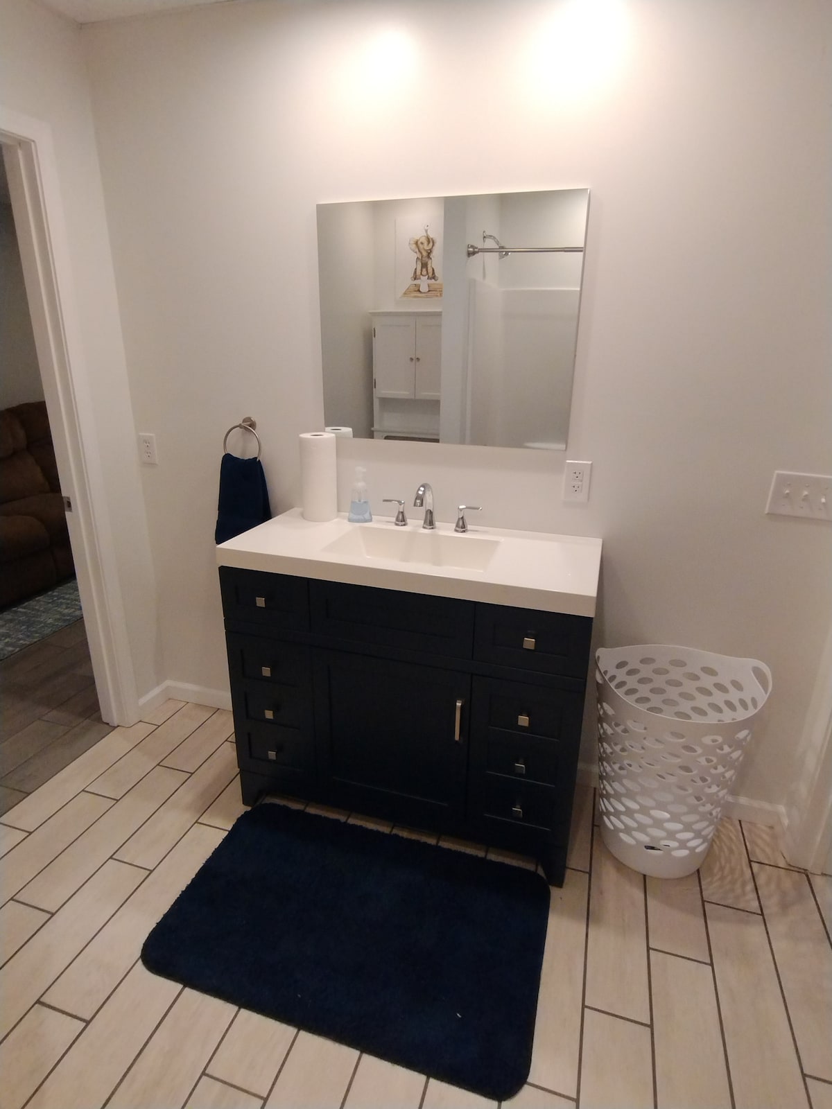 A modern bathroom features a sleek black vanity with dual sinks and a large mirror above. A white laundry basket is placed beside the vanity, and a dark blue bath mat is positioned in front. Neutral-colored tiles line the floor, creating a clean and tidy appearance.