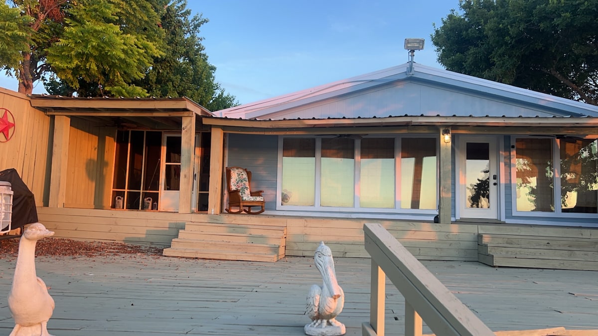 A waterfront house is highlighted by a spacious deck, surrounded by trees and a clear sky. Large glass windows offer views of the water, while a comfortable rocking chair is positioned on the deck. Decorative ducks are visible, enhancing the tranquil outdoor space.