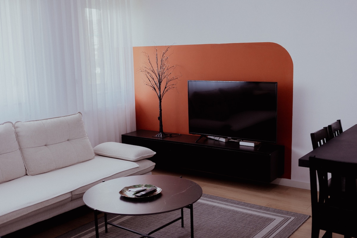 A modern living area is featured, highlighted by a stylish orange accent wall. A flat-screen television is mounted above a sleek black media console. A light-colored sofa is positioned beside a round coffee table, with a simple decorative item resting on it. Natural light filters through sheer curtains.