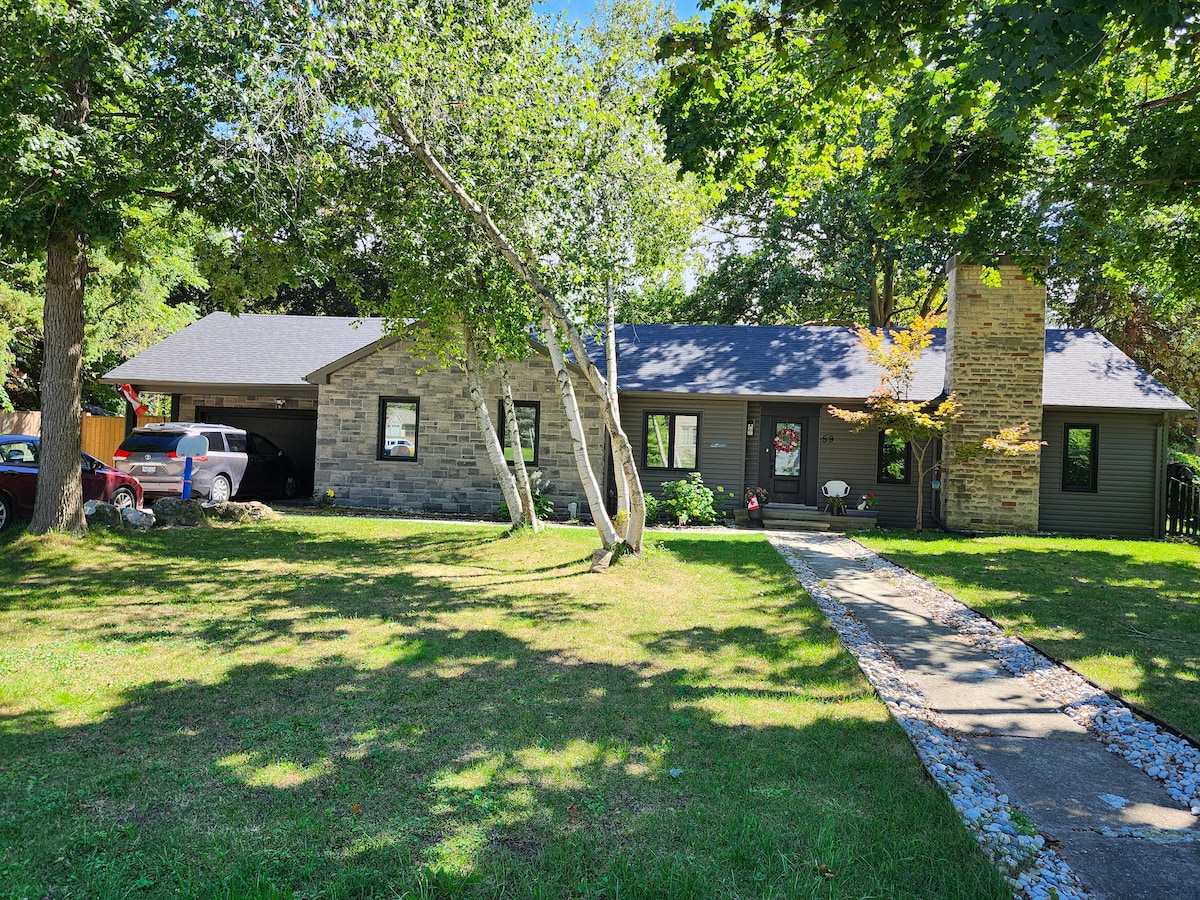 A single-story home is set amidst lush greenery, featuring a stone facade and a chimney. A paved walkway leads to the entrance, bordered by well-maintained grass. Large trees provide shade, contributing to a serene outdoor environment.