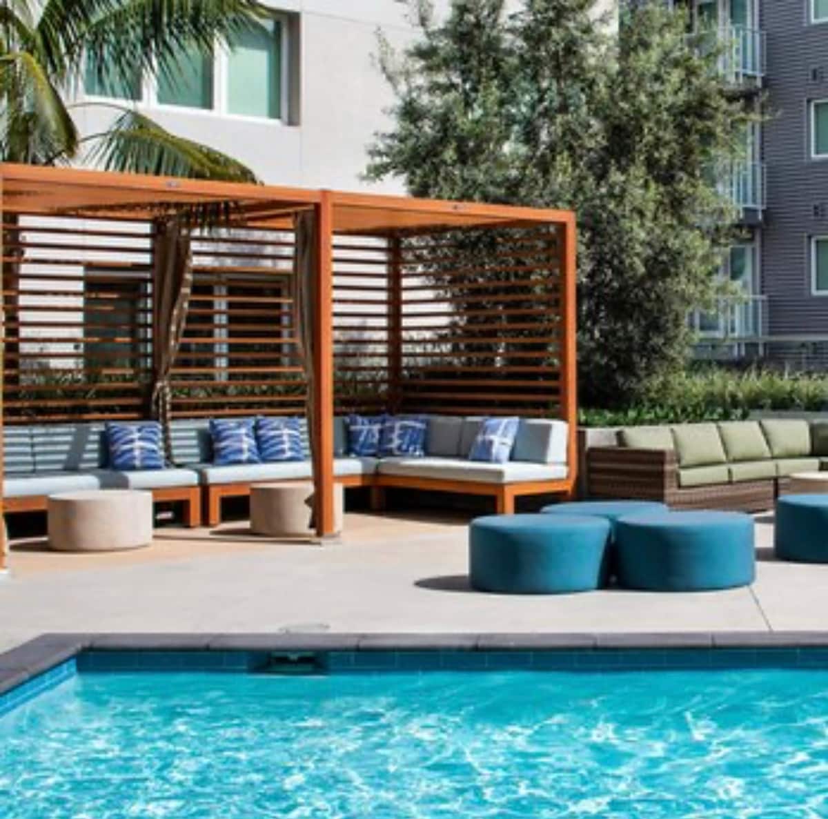 The outdoor area features a refreshing saltwater pool surrounded by comfortable seating arrangements. Canopy-like structures provide shade over lounge areas, while stylish blue ottomans complement the modern furnishings. Lush greenery is visible in the background, enhancing the relaxing atmosphere.