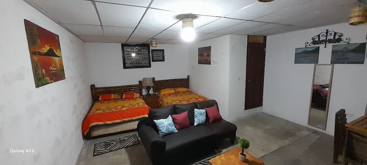 The room features two spacious double beds adorned with colorful bedding, accompanied by a black sofa with decorative cushions. A mirror and a small table are visible, along with framed artwork on the walls, contributing to a cozy and functional living space.