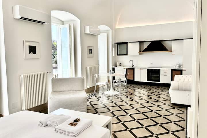 Grand salon de luxe avec cuisine moderne ouverte, équipée d'un four et d'une cuisinière à induction. Table à manger élégante au centre, canapé spacieux et confortable. Lit double inséré dans un environnement harmonieux et raffiné.