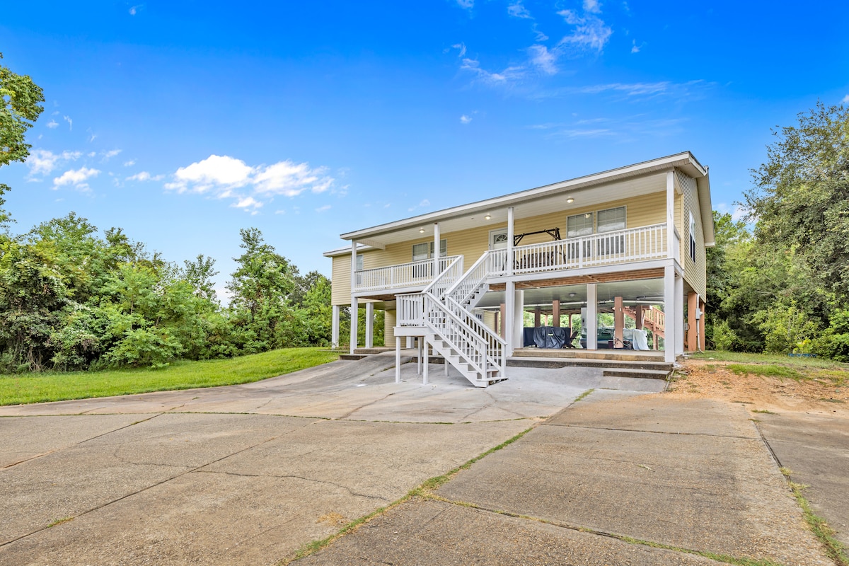A spacious raised house is showcased, featuring a welcoming front staircase leading to a large porch with white railings. Surrounding greenery provides a natural backdrop, while the paved driveway offers ample parking space.