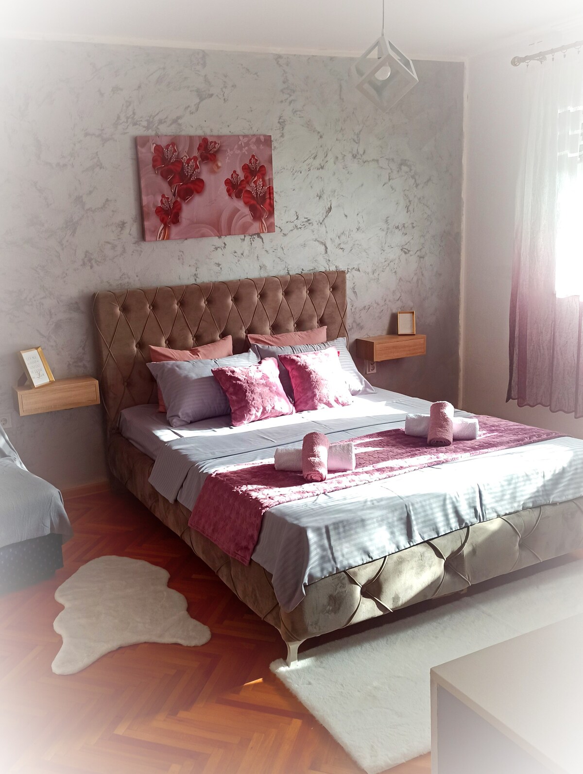 A well-appointed bedroom features a spacious bed with decorative pillows and a textured headboard. Soft pink accents are complemented by light-toned walls and natural light from a window with sheer curtains. A decorative artwork hangs above the bed, and small bedside tables are positioned on either side.