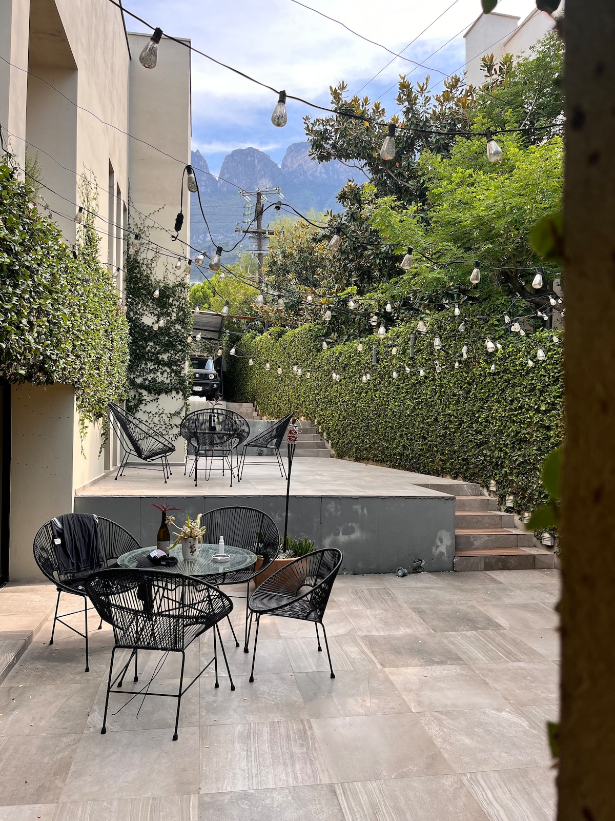 An outdoor area is surrounded by lush greenery, featuring several black wire chairs arranged around a small table. Strands of decorative lights hang overhead, illuminating the space. In the background, distant mountains are visible, enhancing the serene atmosphere of the setting.
