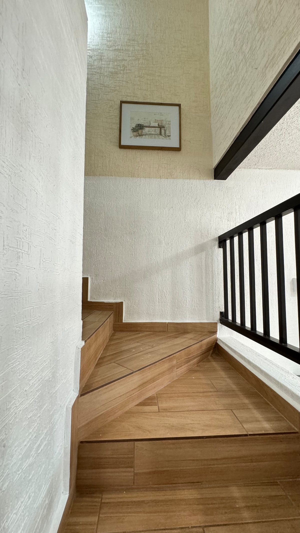 Stairs with wooden steps lead upward, framed by a sleek black railing. Light-colored walls create a bright ambiance, while a framed artwork adorns the wall near the top of the staircase, adding a touch of decor.