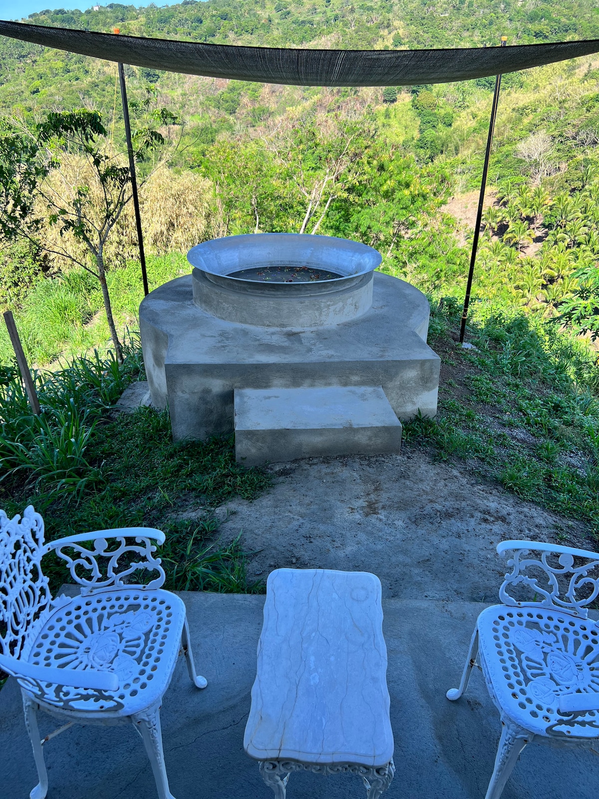 A circular fire ring is set atop a stone base, surrounded by lush greenery. Two white metal chairs with intricate designs are positioned beside a small marble top table, inviting relaxation in the outdoor setting. A shaded canopy protects the area from direct sunlight.