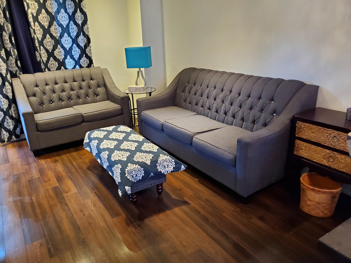A living area features a pair of stylish gray upholstered sofas, one slightly larger, arranged with a decorative ottoman in front. A small side table with a blue lampshade stands beside the seating, with dark curtains framing the window.