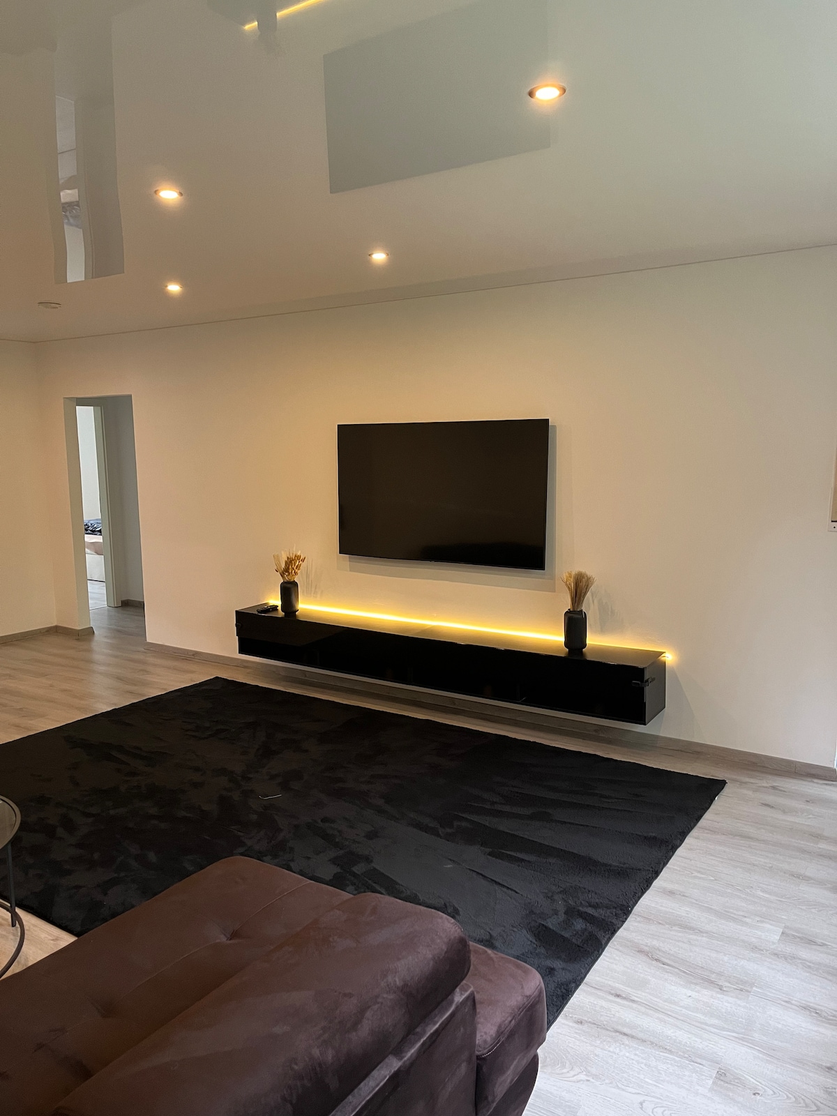 A living room features a mounted television above a sleek, low-profile console. Soft lighting from the shelf creates a gentle glow. A plush, dark area rug extends across the floor, complementing the light wood flooring. A brown couch adds comfort to the space.