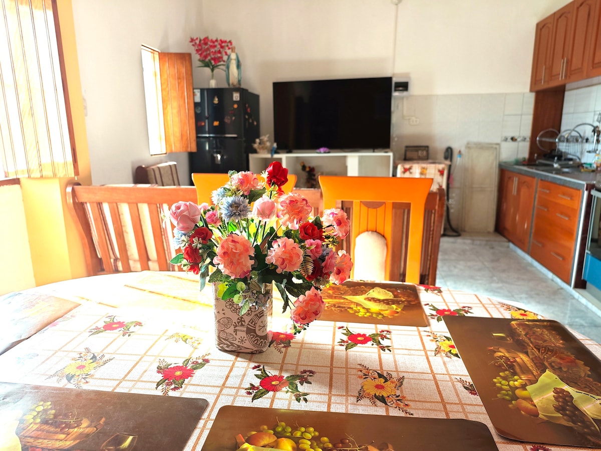 A spacious dining table is adorned with a floral centerpiece, surrounded by wooden chairs. In the background, a large smart TV is positioned near a well-equipped kitchen, featuring wooden cabinetry and essentials for cooking. Natural light filters through the nearby window.