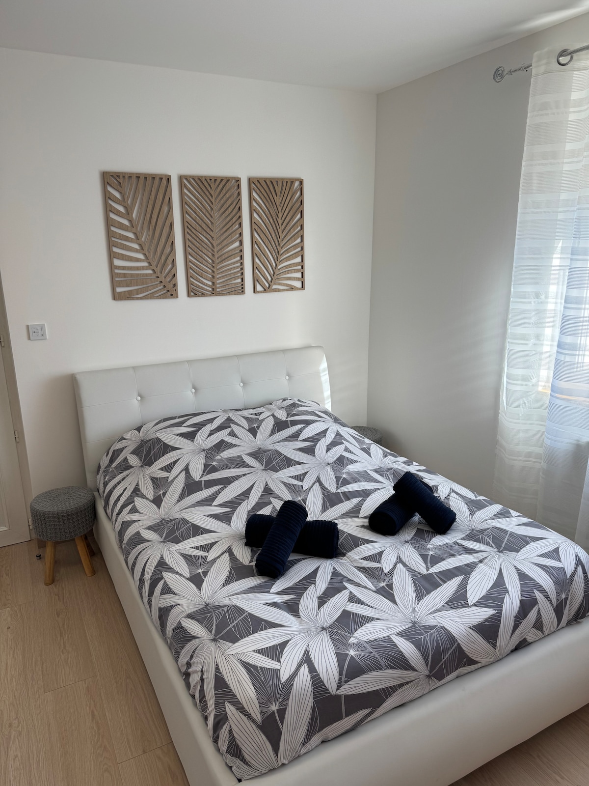 The bedroom features a spacious bed with a botanical patterned duvet cover. Three decorative wall panels are displayed above the bed. Natural light enters through a window adorned with sheer curtains, complemented by a small stool placed beside the bed.