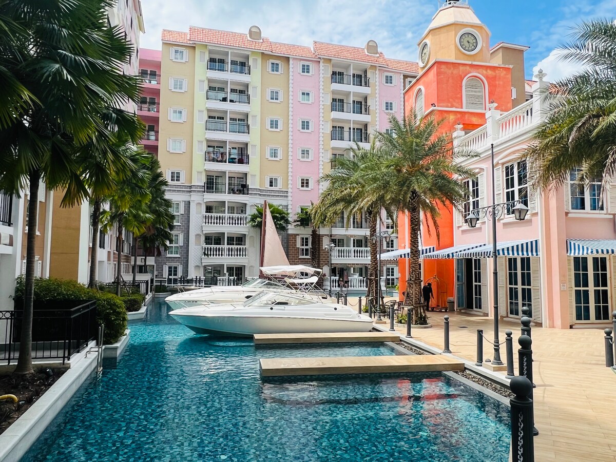 The image showcases a marina-like setting with a sleek white boat moored beside a pool surrounded by lush palm trees. Colorful residential buildings provide a vibrant backdrop, while a charming clock tower adds an architectural element to the scene.